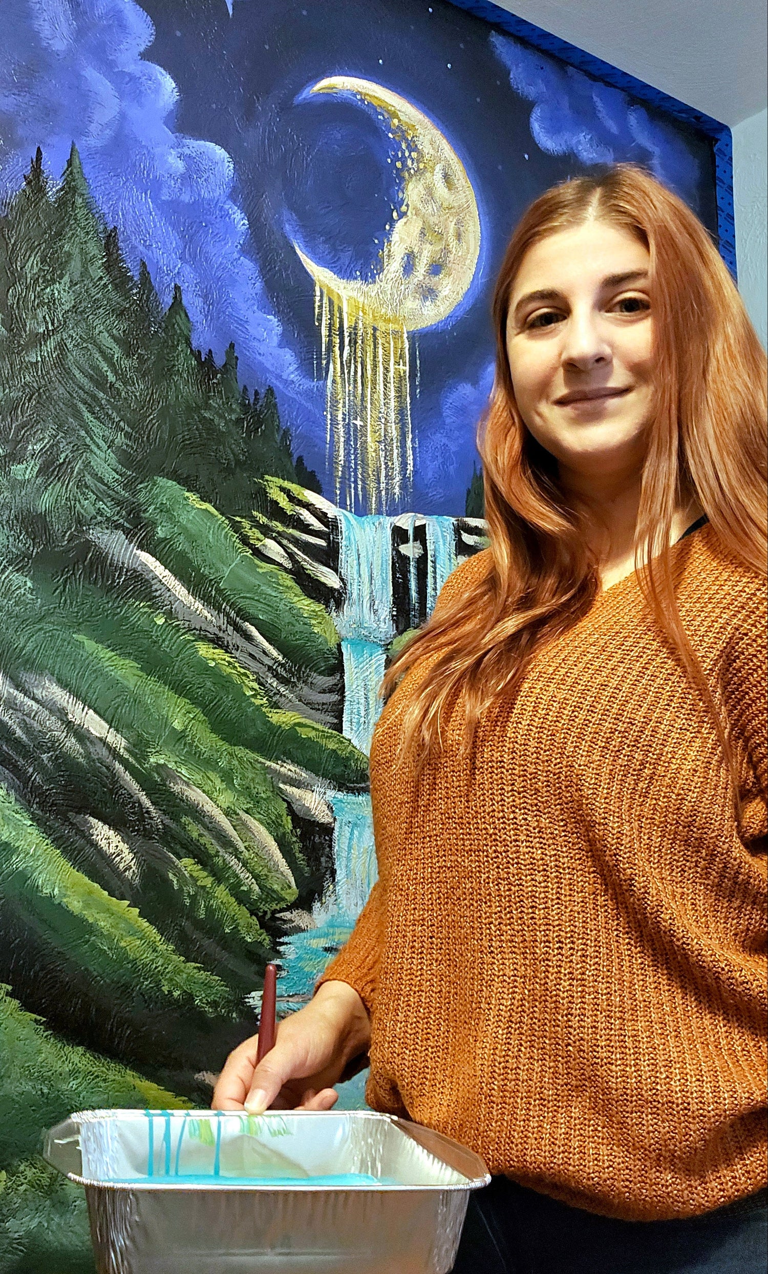 Artist painting a scenic mural with trees and a moonlit sky.